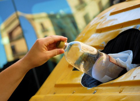 A person's hand is shown dropping a clear plastic bottle into a yellow underfloor container, likely part of a waste collection point on a pavement or driveway. The bottle appears to contain some residual liquid and has a green cap. The container, which has a circular opening, is mounted on a bright yellow surface that is possibly made of metal or plastic, with visible screws securing it in place. In the background, a blurred building or structure with windows can be seen, indicating an urban environment, possibly within Plumstead or nearby areas. This image relates to rubbish collection and waste disposal services provided by Commercial Waste Plumstead, with the focus on proper disposal of plastic bottles as part of responsible waste management.