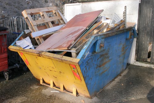 A large industrial-sized skip, painted blue with visible rust and wear, is positioned on a paved surface in an outdoor area, filled with various construction and renovation debris including wooden pallets, laminated boards, and metal fixtures. The skip is situated against a concrete wall, partially shadowed by nearby objects, with some of the waste stacked irregularly, protruding above the top edge. The surrounding environment suggests a commercial or domestic property in Plumstead, with evidence of ongoing waste disposal or clearance activity. A portion of a black wooden fence is visible to the right, and the surface beneath the skip is slightly damp, indicating recent cleaning or weather exposure. This image exemplifies typical rubbish removal tasks managed by Commercial Waste Plumstead, who facilitate the collection and disposal of such waste materials from local premises.