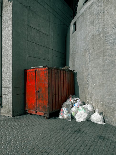 A narrow urban alleyway cluttered with accumulated rubbish and waste materials, featuring a large, black, wheeled rubbish bin in the foreground, positioned on a dusty, uneven asphalt surface. Behind t