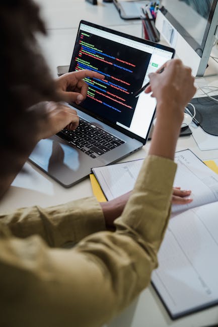 A person seated at a desk working on a laptop, which displays a screen filled with lines of colorful computer code. The individual is holding a pen or stylus in their right hand, pointing towards the 