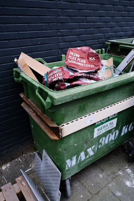 A large green waste bin positioned outdoors on a paved surface against a dark brick wall. The bin is filled with plastic bags of garden or building materials, including a red bag labeled 'FK200 FLEXY 