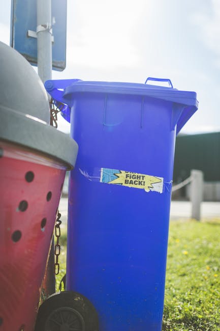 A large green waste bin positioned outdoors on a paved surface against a dark brick wall. The bin is filled with plastic bags of garden or building materials, including a red bag labeled 'FK200 FLEXY 